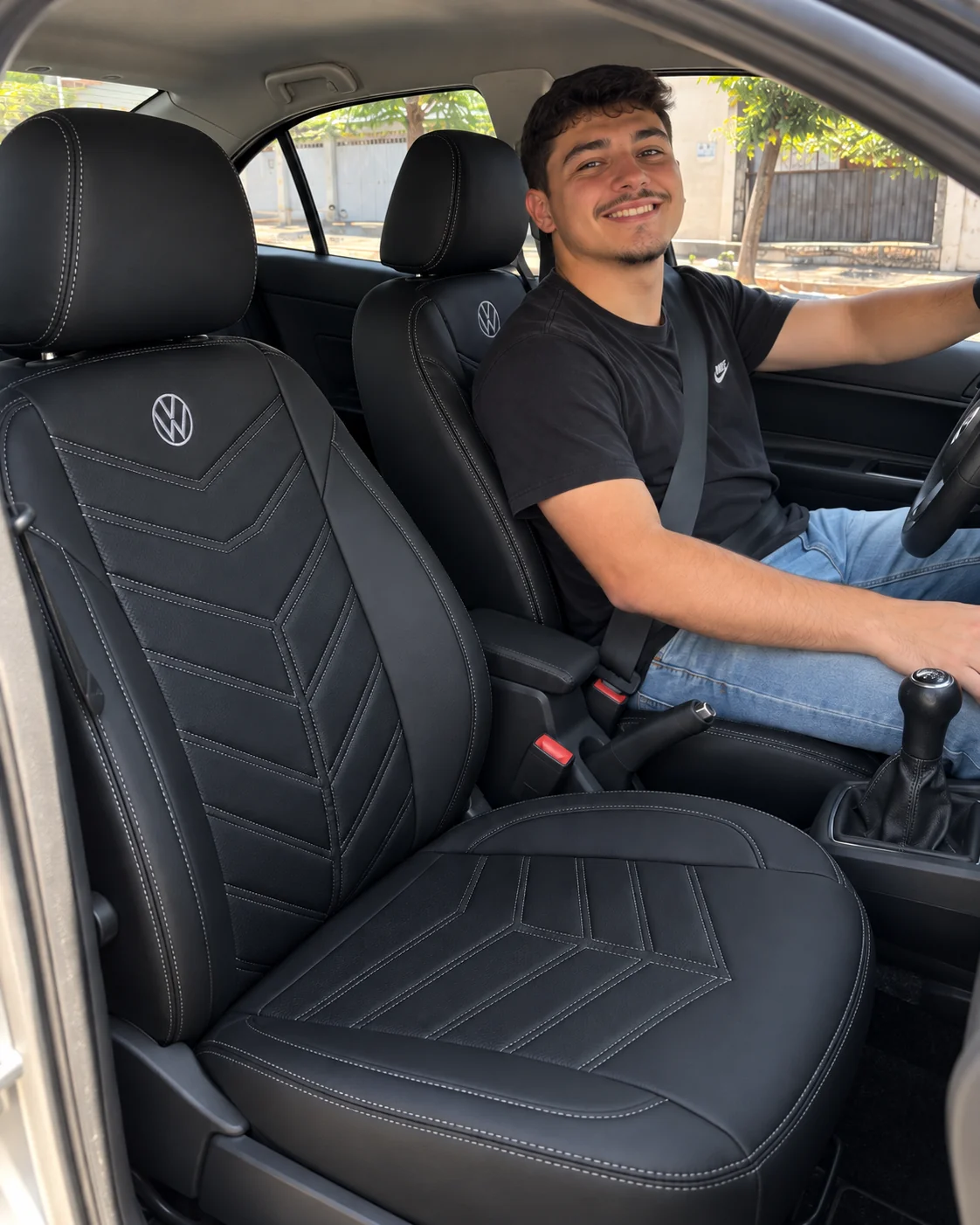 Lucas sorrindo ao volante do Volkswagen Gol com capa Tron preta, linha grafite e logo Volkswagen bordada